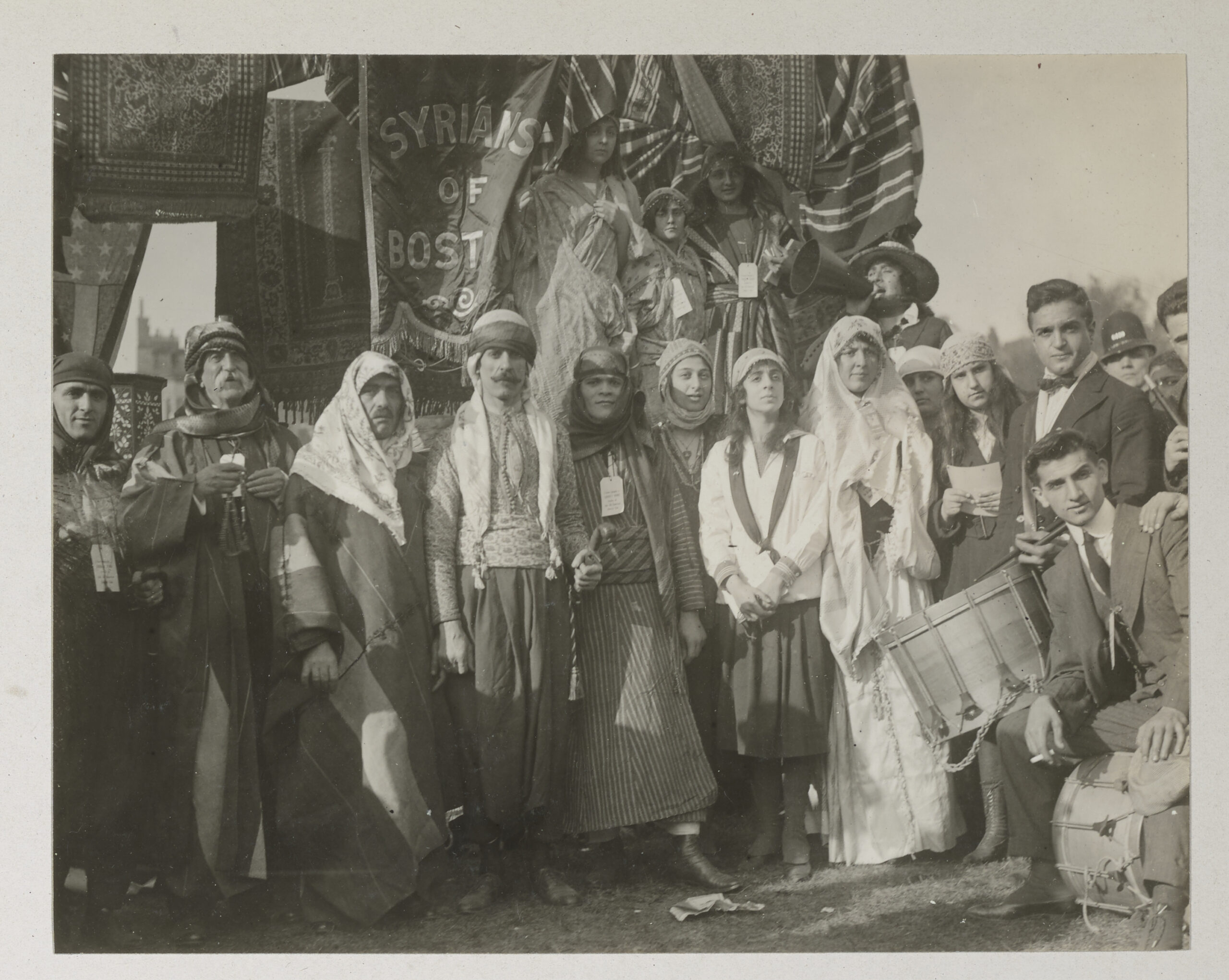 Black-and-white image of a group of Syrians in traditional dress gathered on Boston Common in front of a sign that reads "Syrians of Boston."